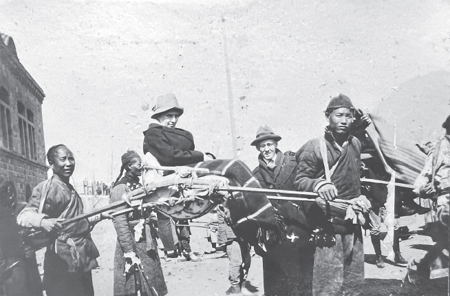 White woman in hat and dark coat being carried in sedan chair by two Chinese men in short dark coats in front of a stone building.