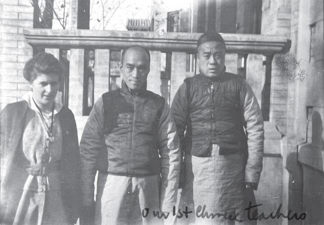 White woman with dark cape and necklace stands with two Chinese men in short dark coats in front of a stone building.