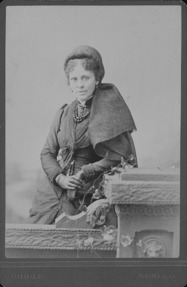 Formal portrait of a woman leaning against a carved stone fence. She wears a dark dress, gloves, a beaded necklace, and a head covering.
