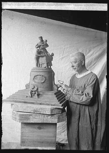 A man by a clay sculpture of a woman holding a baby with children playing around her skirts. The sculpture’s plinth is engraved with the word ‘Mammy.’