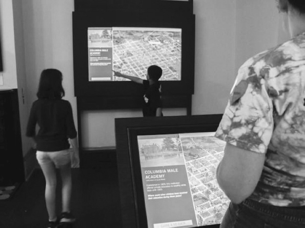 Two digitized maps of Columbia Male Academy. Two children and one adult interact with the images.