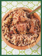A wooden bowl of Hoppin’ John on a patterned tablecloth. There is a spoon sticking out of the bowl.