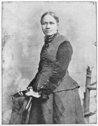 Formal portrait of a woman standing with her hands resting on the back of an upholstered chair. Her hair is pulled back; she wears a dark dress and vest.