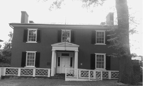 A two-story brick house with white windows, black shutters, and a white column entryway. A white fence and brick pathway are in front.