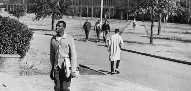 Figure 8. A Black man with books under one arm walks on a stone path. Five people are in the background.