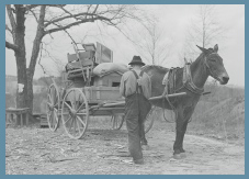 A mule is hitched to a wagon loaded with household items. A man stands facing the wagon.