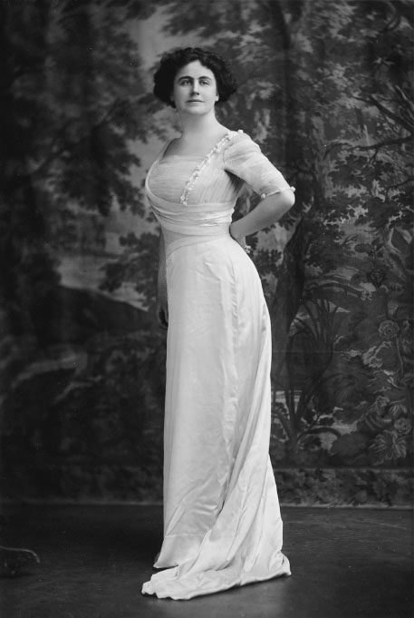 A portrait of a woman standing in a long white dress in front of a woodland backdrop.