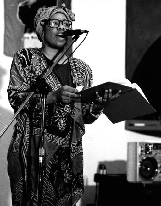A woman, wearing African dress and head cover, stands at a microphone and reads from a book.