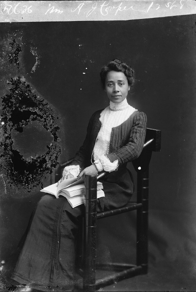Formal portrait of a young woman, seated with an open book in her lap. She wears an elegant full-length dress and looks directly into the camera.