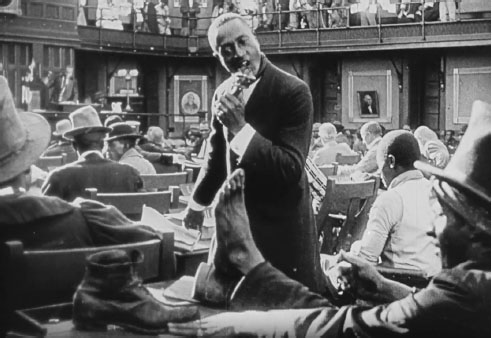A room full of suited men at desks. A man stands and eats a chicken leg, looking at a bare foot on the desk in front of him.